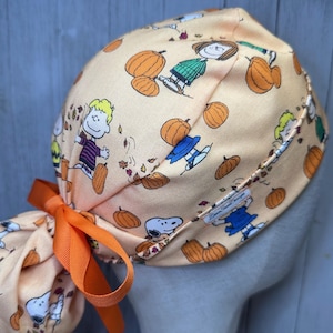 May include: A light orange scrub cap featuring a repeating pattern of cartoon characters, pumpkins, and falling leaves. The cap is tied with an orange ribbon. The design includes characters like Snoopy and Charlie Brown.