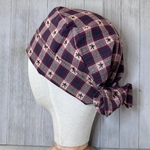 May include: A red and tan plaid bandana with black stars, tied into a bow at the back, on a mannequin head.