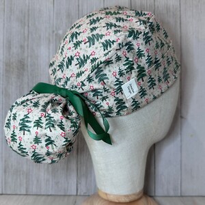 May include: A scrub cap with a Christmas tree pattern in green and red on a cream background. The cap is tied with a green ribbon and has a matching ponytail cover. A small white tag with text is visible.