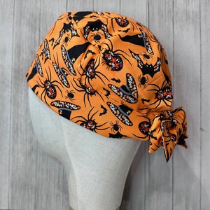 May include: Orange Halloween-themed scrub cap featuring a repeating pattern of black bats, spiders, and witch hats. The cap is tied at the back with a bow. The cap is displayed on a mannequin head.