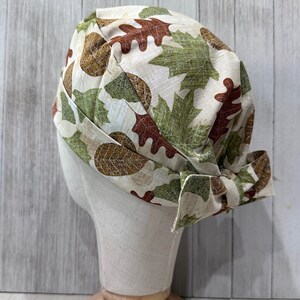 May include: A beige fabric hat with a fall leaf pattern in shades of brown and green. The hat is tied in a bow on the side and is displayed on a mannequin head.