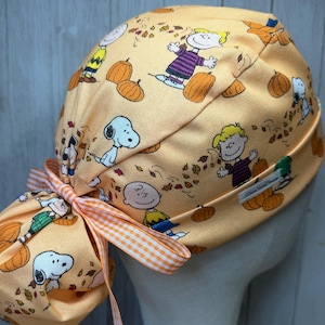 May include: A peach-colored scrub cap featuring a repeating pattern of Peanuts characters including Snoopy, Charlie Brown, and Lucy surrounded by pumpkins and autumn leaves. The cap includes an orange and white gingham ribbon tie at the back.
