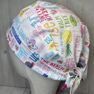 May include: White scrub cap with a beach theme, featuring text and illustrations of flip-flops, pineapples, and beach phrases. The cap is tied at the back with a matching fabric tie.