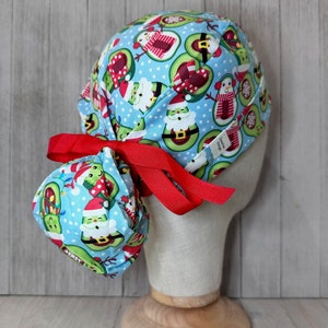 May include: A light blue scrub cap with a festive avocado Christmas print. The cap features avocados dressed as Santa Claus, snowmen, and wrapped in Christmas lights. A red ribbon is tied in a bow at the back. The cap is on a mannequin head.