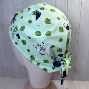 May include: A light green surgical scrub cap with a repeating pattern of cartoon characters and green shamrocks. The cap is tied at the back and is displayed on a mannequin head. The fabric has a light green background.