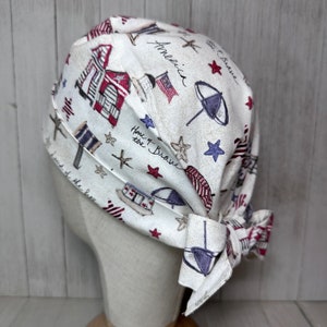 May include: A white fabric surgical cap with a red, white and blue pattern. The pattern includes stars, stripes and images of beach umbrellas, beach houses and boats. The text "America, Home of the Brave, Land of the Free" is printed on the cap.