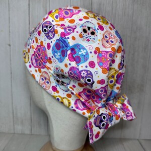 May include: White surgical scrub hat with a colourful pattern of sugar skulls, pink, purple, blue, orange and yellow.