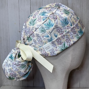 May include: A yellow surgical scrub hat with a floral print featuring a yucca plant design. The hat has a long tie that can be tied in a knot at the back of the head. The hat is made of a soft, comfortable fabric and is perfect for nurses, doctors, and other healthcare professionals.