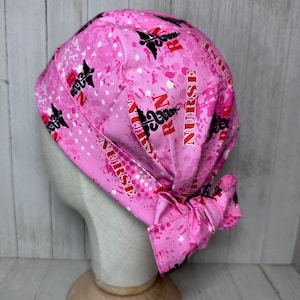 May include: Pink scrub hat with a white and black splatter pattern. The hat has the words "RN" and "NURSE" repeated in red and black. The hat is tied at the back.