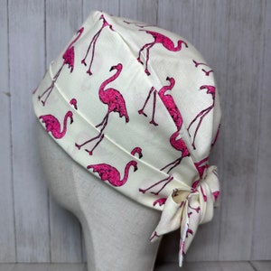 Puede incluir: Un gorro de quirófano blanco con estampado de flamencos rosas. El gorro está atado en la parte posterior con un lazo.