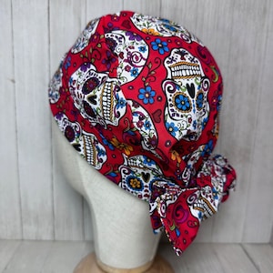 May include: Red surgical scrub hat with a colorful sugar skull pattern. The hat has a tie at the back.