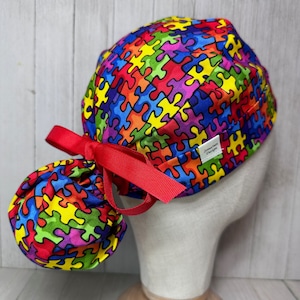 May include: A red, blue, yellow, green, and purple puzzle piece patterned scrub hat with a red tie. The hat has a small white label that says "Jenna Rae Designs".