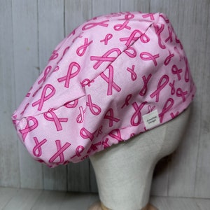 May include: Pink scrub hat with a pink ribbon pattern. The hat has a small white tag that says "Zenadan Design".