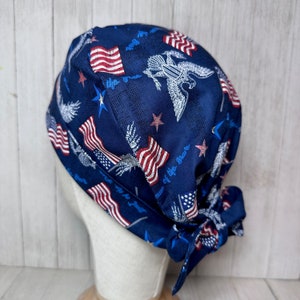 May include: A navy blue scrub cap with a patriotic design featuring American flags, stars, and eagles. The cap is tied at the back and has the phrase "Home of the Brave" in a script font. The fabric has a textured appearance.
