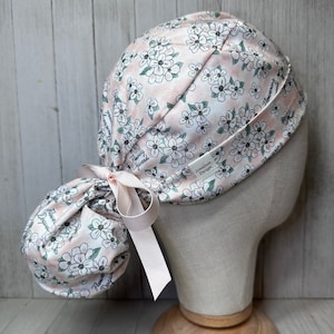 Peut inclure: Un bonnet de travail chirurgical rose et blanc à fleurs avec un trou pour la queue de cheval et un ruban rose. Le bonnet porte l'inscription "Flowering Goodness" et "ZonaSun Designs".