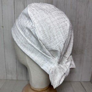 May include: A light grey fabric scrub hat with a bow at the back. The hat has a textured pattern.