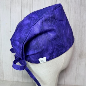 May include: Purple scrub hat with a tie-back design. The hat has a white label with the text "Zenith Design".