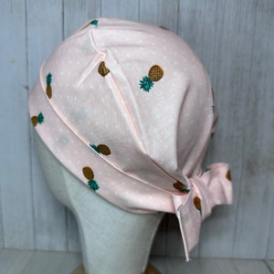 May include: A pink surgical scrub hat with a white polka dot pattern and a pineapple print. The hat is tied at the back.