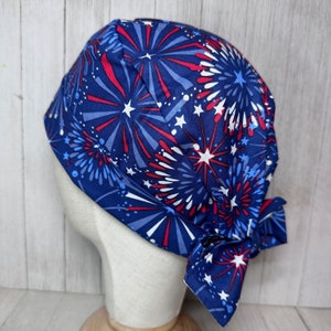 May include: A blue, red and white patterned bandana-style surgical cap with a fireworks design. The cap has a tie at the back.