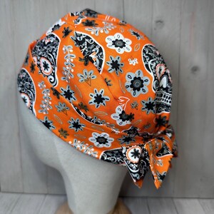 May include: Orange and black skull and floral print fabric scrub hat. The hat is tied in a bow at the back.