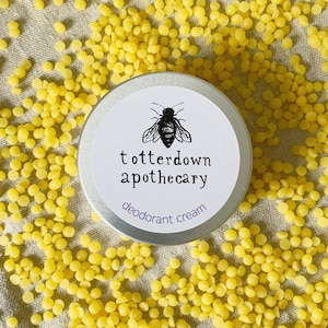 May include: A silver tin with a white label that says "totterdown apothecary" and "deodorant cream". A black and white illustration of a bee is on the label. The tin is surrounded by yellow beeswax pellets.