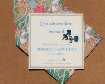 May include: A pair of blue glass stud earrings made from recycled Bombay Sapphire gin bottles. The earrings are displayed on a card with the text "Gin-dependent woman!" and "Made from recycled BOMBAY 'SAPPHIRE' Gin bottles."