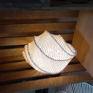 May include: A white, woven, spiral-shaped lampshade with a warm light shining through it. The lampshade is sitting on a wooden shelf.