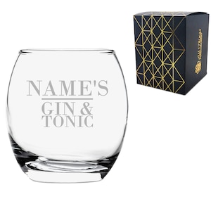 Op de afbeelding: Een helder glazen tumbler met de tekst "NAME'S GIN & TONIC" gegraveerd aan de zijkant. Het glas is verpakt in een zwarte doos met een gouden geometrisch patroon.