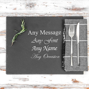Könnte beinhalten: Ein schwarzes Schiefer-Platzset mit einer weißen Aufschrift, die "Any Message Any Font Any Name Any Occasion" lautet. Eine graue Serviette mit weißen Streifen liegt rechts auf dem Platzset, darauf liegen eine silberne Gabel und ein Messer.