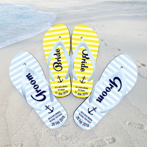 Puede incluir: Un par de chanclas rayadas azules y amarillas con la palabra "Groom" impresa en una y la palabra "Bride" impresa en la otra. Las chanclas tienen un símbolo de ancla y el texto "Al & Justin's Wedding, Port Douglas, 11 June 2019" impreso en la parte inferior.