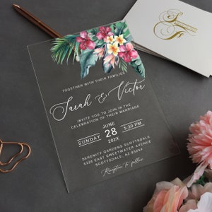 Pode incluir: Um convite de casamento em acrílico transparente com desenhos florais tropicais em tons de rosa, verde e amarelo. O convite apresenta o texto "Sarah & Victor" e os detalhes da sua cerimônia de casamento no domingo, 28 de junho de 202X às 17h30 no Serenity Gardens Scottsdale, 5326 East Sweetwater Avenue, Scottsdale, AZ 85254. O convite também inclui o texto "Recepção a seguir."