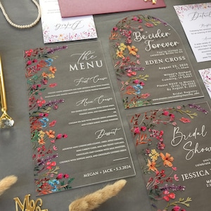 Könnte beinhalten: Set aus transparentem Acryl-Hochzeitspapeterie mit floralen Designs. Enthält eine Menükarte, eine Einladung und eine Detailkarte. Die Menükarte listet die Gänge auf, und die Einladung enthält Eventdetails. Die floralen Akzente zeigen rosa, orange und blaue Blumen.