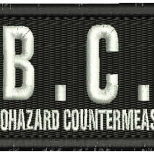 May include: Black patch with white embroidered text "U.B.C.S." and the words "Umbrella Biohazard Countermeasure Service" below.