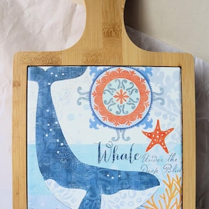 Peut inclure: Planche de service en bambou avec un carreau carré représentant une illustration de baleine bleue. Le carreau comprend le texte "Whale Under the Deep Blue" avec des accents d'étoile de mer et de corail. La planche a une poignée.