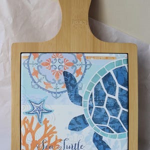 May include: A bamboo chopping board with a decorative tile insert featuring a blue sea turtle design. The tile includes a starfish, coral, and the words "Sea Turtle". The board has a handle.
