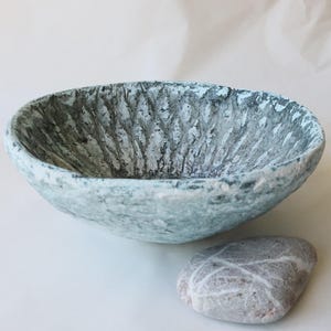 Peut inclure: Un bol rond et peu profond avec une surface texturée et irrégulière présentant une finition grise et blanche tachetée. L'intérieur présente des rainures verticales profondes. À côté du bol se trouve une pierre grise ovale et lisse.