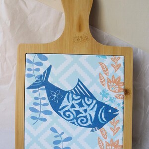 Può includere: Tagliere decorativo in legno con manico. Il centro presenta un disegno di pesce blu su uno sfondo a motivi blu chiaro e bianco. Accenti floreali arancioni e blu circondano il pesce. Il tagliere è in bambù.