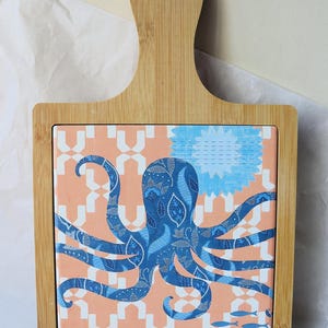 May include: A bamboo cutting board with a square tile featuring a blue octopus design on a peach and white patterned background. The octopus has intricate details, and the board has a handle.