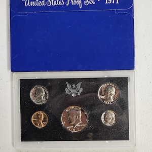 May include: A 1971 United States Proof Set in a blue cardboard sleeve with white text. The clear plastic case contains five coins: a Lincoln cent, a Jefferson nickel, a Roosevelt dime, a Washington quarter, and a Kennedy half dollar, plus an eagle emblem.