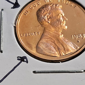 Puede incluir: Una moneda de un centavo estadounidense de color cobre con las palabras "In God We Trust" y "Liberty" inscritas en ella. El año 1983 también es visible en la moneda.
