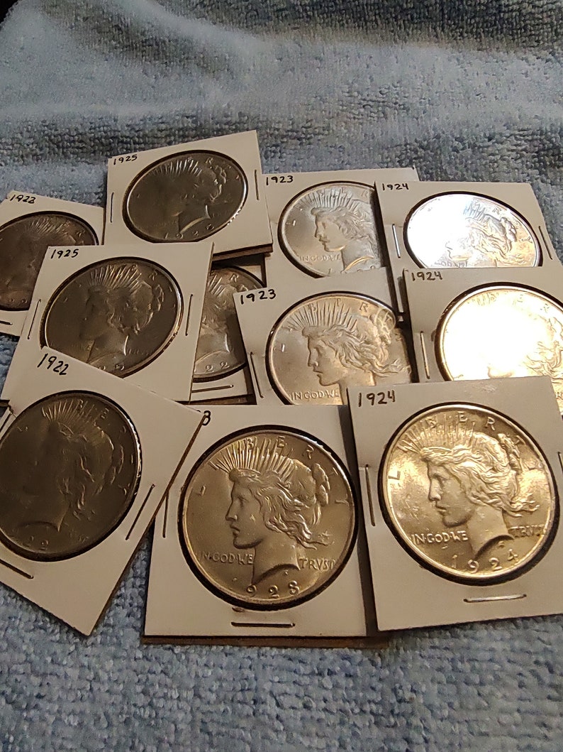 May include: A collection of silver Peace Dollar coins from the 1920s, each coin is in a white cardboard holder with the year of minting printed on it.