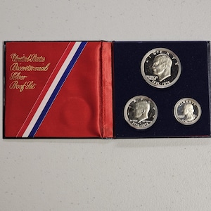 May include: A United States Bicentennial Silver Proof Set in a black display case. The left side features red fabric with a diagonal red, white, and blue stripe and the text United States Bicentennial Silver Proof Set. Three silver coins are displayed.
