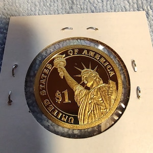 May include: A gold-colored coin with the image of the Statue of Liberty and the text "United States of America" and "$1" on it.
