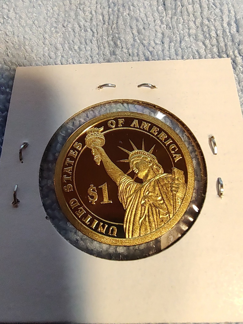 May include: A gold-colored coin with the image of the Statue of Liberty and the text "United States of America" and "$1" on it.
