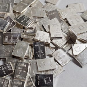 May include: A collection of small, rectangular silver bars scattered across a surface. Each bar has a stamped design, with some featuring the text "999.0 silver" and "valcambi". The bars are a bright, reflective silver color.