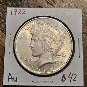 May include: A silver 1922 Peace Dollar coin with the inscription "IN GOD WE TRUST" and "LIBERTY" on the obverse. The coin is in a white cardboard holder with the year "1922" and the price "$42" written on it.