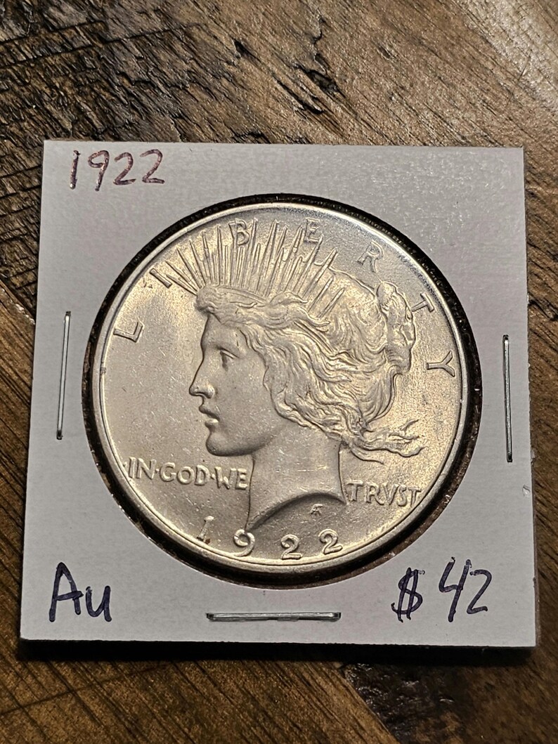 May include: A silver 1922 Peace Dollar coin with the inscription "IN GOD WE TRUST" and "LIBERTY" on the obverse. The coin is in a white cardboard holder with the year "1922" and the price "$42" written on it.