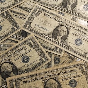 May include: A collection of vintage one-dollar bills, featuring a portrait of George Washington. The bills are tan with black ink, and the text "ONE DOLLAR" is prominently displayed. The bills are from the 1957 series.