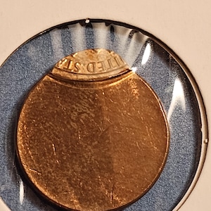 May include: A copper-colored coin, partially visible, encased in a clear plastic holder. The coin's edge shows the words "UNITED STATES" in raised lettering. The holder is set against a blue background.