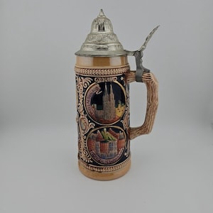 May include: A ceramic beer stein with a metallic hinged lid and a textured handle. The body features two circular relief panels depicting the Cologne Cathedral with the text KÖLN RHEIN DOM and the Munich Hofbräuhaus with the text MÜNCHEN HOFBRÄUHAUS.